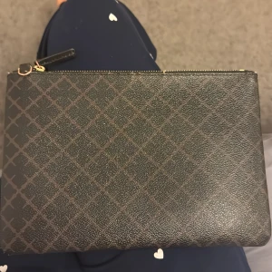 Brun kuvertväska från Malene birger - Snygg brun kuvertväska i skinnimitation med rutigt mönster och guldfärgad dragkedja. Väskan har en enkel och stilren design, perfekt att bära i handen eller lägga i en större väska. Insidan är fodrad i beige tyg. ( fläckar finns inuti väskan, se bild) 