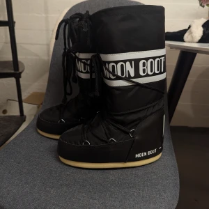 Svarta Moon Boots i nyskick - Svarta Moon Boots med vit logga och snörning framtill. Skorna har en hög och vadderad design med platt sula och är perfekta för kalla vinterdagar. Skorna är i mycket bra skick, strl 35-38.