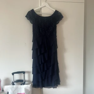 Marinblå långklänning med spets - Säljer en marinblå långklänning med vacker spets upptill och glittriga paljetter. Klänningen har korta ärmar och flera lager volanger nedtill som ger en snygg och flödande look. Perfekt för dig som vill sticka ut med elegans och stil. Aldrig använd.