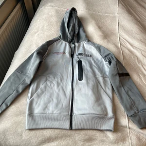 Grå hoodie från Hoodrich med dragkedja - Snygg grå hoodie från Hoodrich med svarta detaljer och hel dragkedja framtill. Tröjan har huva, långa ärmar och en bröstficka med dragkedja. Perfekt för en streetwear-look och chill dagar.