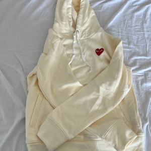 Beige hoodie Comme des Garçons Play - Säljer en Beige hoodie från Comme des Garçons Play med det ikoniska röda hjärtat broderat på bröstet. Tröjan har huva med snörning, känguruficka och är tillverkad i mjuk bomull. Perfekt för dig som gillar clean streetstyle med en twist.