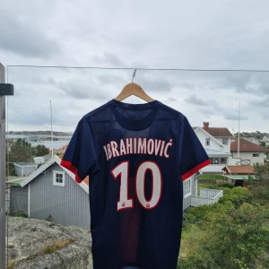 PSG Ibrahimović matchtröja Nike - Snygg PSG fotbollströja med Ibrahimović och nummer 10 på ryggen. Tröjan är marinblå med röd och vit detalj framtill, klassisk rund hals och korta ärmar. Materialet är lätt och ventilerande polyester, perfekt för träning eller match. PSG-logga och Nike-märke på bröstet.