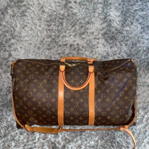 Louis Vuitton *Band Keepall 55 Monogram resväska - Säljer en klassisk Louis Vuitton Keepall resväska i brunt monogrammönster med ljusbruna skinnhandtag och axelrem. Väskan har dragkedja upptill och detaljer i äkta skinn. Perfekt för weekendresor och har ikoniska LV-detaljer.