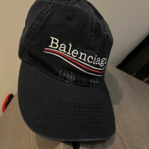 Mörkblå keps från Balenciaga - Snygg mörkblå keps från Balenciaga med broderad logga framtill och röda samt vita vågiga linjer under texten. Klassisk böjd skärm och justerbar passform. Tillverkad i bomull för en bekväm känsla och stilren look. Fett skön keps säljer pga att den inte kommer till användning ny skick.