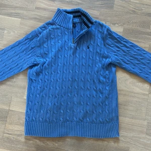 Blå stickad tröja Polo Ralph Lauren - Säljer en blå stickad tröja från Polo Ralph Lauren med kabelstickat mönster och half zip i halsen. Tröjan har ribbad krage med mörkblå ränder, långärmad design och klassisk Polo-logga broderad på bröstet. Perfekt för dig som gillar stilren och sportig look.