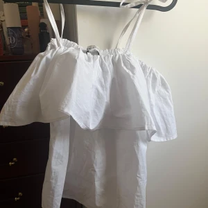 Vit volangig linnetopp med smala band - Supersöt vit linnetopp med smala axelband och volang över bysten. Toppen har en luftig och flowy känsla, perfekt för varma dagar. Resår upptill ger en snygg passform och den är lätt att styla till jeans eller kjol.