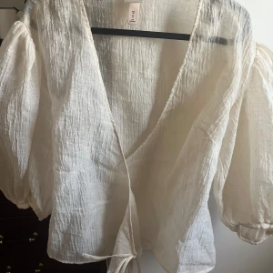 Vit omlottblus med puffärm - Säljer en superfin vit omlottblus i luftig och lätt struktur. Blusen har puffiga trekvartsärmar och knytband i midjan. Perfekt för dig som gillar en romantisk och trendig look. Passar snyggt till både jeans och kjol.
