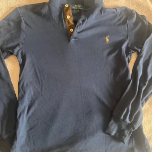 Marinblå långärmad pikétröja Polo Ralph Lauren - Snygg marinblå långärmad pikétröja från Polo Ralph Lauren med klassisk krage och knappar framtill. Ikoniska broderade loggan i gult på bröstet. Mjuk bomullskvalitet som är skön att bära. Perfekt till jeans eller chinos för en clean look. Perfect för en stilig och grisch look. Köpt på 899kr.