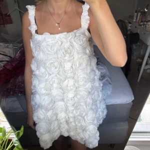 Vit kort klänning med rosor - Supersöt vit kort klänning med 3D-rosor över hela tyget och breda axelband. Klänningen har en fluffig och voluminös form som ger en unik look. Perfekt för dig som vill sticka ut och älskar detaljer. Materialet känns mjukt och har struktur tack vare rosapplikationerna. Från nakd 