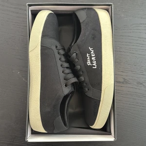 Svarta Saint Laurent sneakers strl 43 - Svarta sneakers från Saint Laurent med vit sula och handskriven logga på sidan. Skorna har snörning, rund tå och är tillverkade i canvas och skinn. Klassisk och stilren modell med kontrasterande detaljer och diskret brand på innersulan. Skicka meddelande vid intresse! 