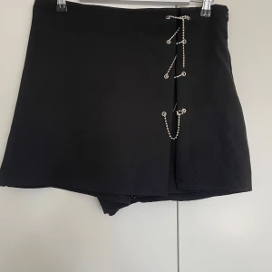 Svart skorts med kedje-diamant detaljer - Svarta skorts med coola silverfärgade kedjor som går snett över framsidan och fästs i metallöglor. Kjolen har en rak passform och är gjord i ett slätt syntetmaterial. Perfekt för dig som gillar edgy detaljer och vill sticka ut lite extra.