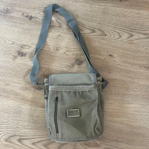 Olivgrön axelväska från BP World - Säljer en olivgrön axelväska från BP World med justerbar rem. Vintage väska som passar både tjejer och killar. Den är i använt skick w lite slitningar men själv tycker jag bara det är nice detalj.