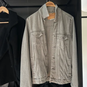 Grå jeansjacka från Pull&Bear - Säljer en grå jeansjacka från Pull&Bear i klassisk modell med knappar framtill och två bröstfickor med lock. Jackan har en avslappnad passform och är tillverkad i slitstarkt jeanstyg. Perfekt att slänga över en hoodie eller t-shirt för en chill look.