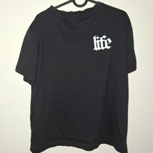 Svart t-shirt med tryck 'life' - Svart t-shirt med vit text 'life' i gotisk stil på bröstet och motiverande citat på ryggen. Rund hals och korta ärmar. Materialet är mjukt och består av 50% bomull och 50% polyester. Passar dig som gillar streetwear och tydliga budskap.
