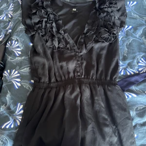 Svart jumpsuit med volang från H&M - Supersnygg svart jumpsuit från H&M med volangdetaljer längs v-ringningen och knappar framtill. Klänningen har resår i midjan för en smickrande passform och är tillverkad i ett lätt, glansigt material som känns lyxigt.
