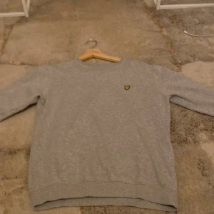 Grå sweatshirt från Lyle & Scott - En stilren grå sweatshirt från Lyle & Scott med klassisk rund hals och det ikoniska gula örnloggan broderad på bröstet. Tröjan har lång ärm och är tillverkad i mjuk bomull, perfekt för en chill och enkel look, tröjan är aldrig använd och är i nytt skick.