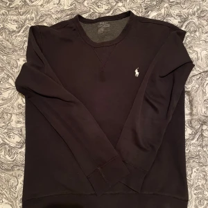 Tröja  Ralph Lauren - Klassisk svart sweatshirt från Polo Ralph Lauren med rund halsringning och vit broderad logga på bröstet. Tröjan har långa ärmar och ribbade muddar vid ärmslut och nederkant. Perfekt för en stilren och avslappnad look. En parfym fläck i mitten. Tvätten går det bort om det ej gör de ja gör retur 