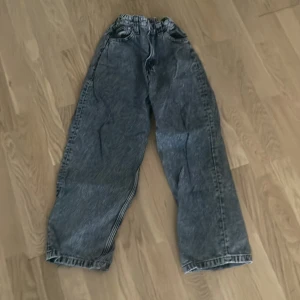 Grå baggy jeans med tvättad look - Säljer ett par grå jeans med tvättad effekt och riktigt baggy passform. De har klassiska fem fickor och breda ben, perfekt för en avslappnad streetstil. Materialet är jeans och färgen är mörkgrå med slitningar för en cool vintagekänsla.