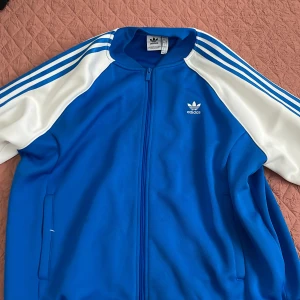 Blå och vit Adidas kofta L - Säljer en klassisk Adidas kofta i blått med vita ärmar och de ikoniska tre ränderna längs axlarna. Jackan har dragkedja framtill, ribbade muddar och Adidas-logga på bröstet. Perfekt för en sportig och avslappnad stil.