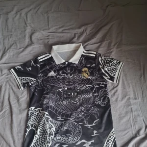 Real Madrid x Adidas svart draktröja - Säljer en svart Real Madrid fotbollströja från Adidas med grymt drakmotiv över hela tröjan. Tröjan har vit krage, vita detaljer och klubbmärke i guld på bröstet. Materialet är lätt och andas, perfekt för match eller träning.