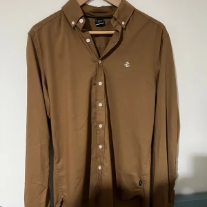 Brun skjorta från Blakely, Small - Snygg brun skjorta från Blakely i storlek Small. Skjortan har klassisk krage, vita knappar och ett broderat ankare på bröstet. Tillverkad i mjuk bomull och har lång ärm. Perfekt för dig som gillar stilrena plagg med diskreta detaljer. Öppen för bundles och frågor!  Man, kille, grisch, Stockholm, plagg, billigt, small, s, stil, stiligt, style, jacka, tröja, skjorta, kvalitet, märke, märkeskläder