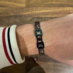 Louis Vuitton brunt armband med monogram - Snyggt armband från Louis Vuitton i brunt med klassiskt monogram och svarta detaljer. Armbandet har metallspänne med LV-logga och texten 'Louis Vuitton'. Perfekt accessoar för att lyfta din stil med en lyxig touch.