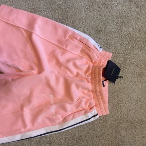 Palm Angels rosa mjukisbyxor - Snygga rosa mjukisbyxor från Palm Angels med vita och svarta ränder längs sidorna. Byxorna har elastisk midja och Palm Angels-logga broderad på benet. Perfekta för en chill och trendig streetwear-look.