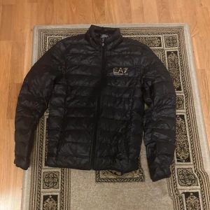 Svart dunjacka från EA7 Emporio Armani - Svart dunjacka från EA7 Emporio Armani med guldfärgad logga på bröstet. Jackan har quiltad design, dragkedja framtill och hög krage. Perfekt för kyliga dagar och riktigt snygg till streetwear-stilen.