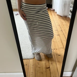 Randig långkjol från Viva Couture - Supersnygg långkjol från Viva Couture i vitt med svarta horisontella ränder. Kjolen är ribbad och har en slits där bak. Den har ett litet hål där bak vid slitsen men det syns knappt när man har på sig. Har ett märke på mitt spegel det är ingen defekt på framsidan!