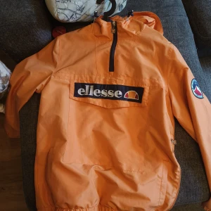 Orange vindjacka från Ellesse M - Snygg orange vindjacka från Ellesse med stor logga på bröstet och patch på ärmen. Jackan har huva, halv dragkedja och stor ficka framtill. Tillverkad i 100% polyester och perfekt för dig som gillar sportig streetstyle.