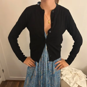 Svart kofta från H&M i bomull XS - En svart, klassisk kofta från H&M i storlek XS. Koftan har rund halsringning, långa ärmar och knäppning framtill med svarta knappar. Tillverkad i mjuk bomull, perfekt att slänga över en klänning eller topp.