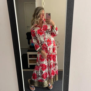 Vit långklänning med röda blommor - Superfin långklänning i vit bas med stora röda blommor och gröna blad. Klänningen har långa ärmar, knappar framtill och coola detaljer i beige och svart mönster vid ärmslut och nederkant. Luftig och flowy modell, perfekt för sommarens alla tillfällen.helt ny