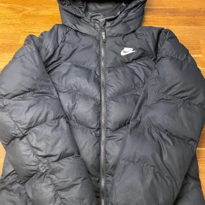 Svart pufferjacka från Nike  - Säljer en svart pufferjacka från Nike i storlek XL. Jackan har huva, dragkedja framtill och klassisk Nike-logga på bröstet. Perfekt för kalla dagar med sin vadderade design och stilrena look. Passar dig som gillar sportig och enkel stil.