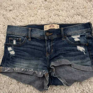 Lågmidjade jeansshorts från Hollister - Snygga blå jeansshorts från Hollister med låg midja och råa, fransiga kanter. Shortsen har slitna detaljer framtill och bakfickor med dekorativa sömmar. Perfekta för sommaren och ger en avslappnad vibe.