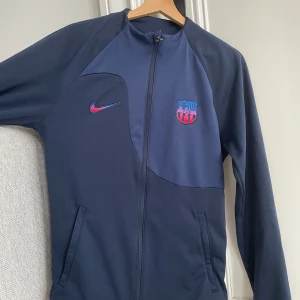 Barça Nike träningsjacka slim fit S - Snygg mörkblå Nike träningsjacka med FC Barcelona-logga och broderad swoosh i rött och blått. Jackan har hel dragkedja, två fickor och är i slim fit-modell. Perfekt för dig som älskar fotboll och vill representera Barça med stil.