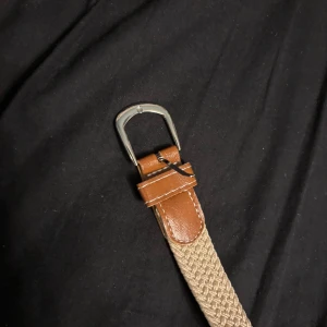 Beige flätat bälte med skinn-detaljer - Snyggt beige bälte med flätad design och bruna skinn-detaljer vid spännet. Spännet är i silverfärgad metall och bältet har en stilren look som passar till många outfits. Perfekt accessoar för att lyfta din stil.