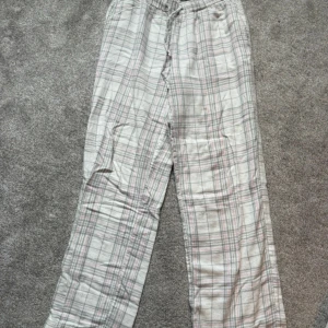 Rutig pyjamasbyxor från BikBok - Supermysiga pyjamasbyxor från BikBok i storlek XS. Byxorna är vita med grått och rosa rutmönster, har resår och snörning i midjan samt raka ben. Finns en liten fläck på de men inte jätte synlig🥰