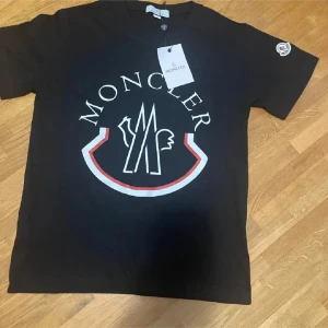 Svart Moncler t-shirt med stor logga - Svart t-shirt från Moncler med stor vit och röd logga framtill och patch på ärmen. Klassisk rund halsringning och korta ärmar. Tillverkad i mjuk bomull för skön känsla och snygg passform. Perfekt för dig som gillar streetstyle och premiumkänsla.
