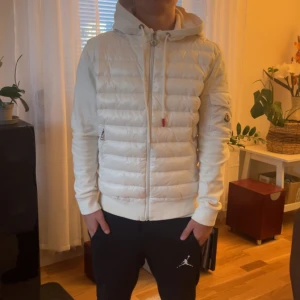 Vit Moncler cardigan med huva - Snygg vit cardigan från Moncler med huva och dragkedja framtill. Jackan har quiltad framsida, släta ärmar och två fickor med dragkedja. Klassisk Moncler-logga på ärmen och snörning i huvan. Perfekt för dig som vill ha en clean och stilren look. Kan sänka pris vid snabb affär fraktar samma dag som köpt. Storlek: S passar 165-180
