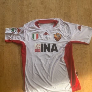 AS Roma Totti #10 bortatröja Kappa - Säljer en klassisk AS Roma bortatröja från Kappa med Totti och nummer 10 på ryggen. Tröjan är vit med röda detaljer på sidor och krage, samt klubbmärke och sponsortryck på bröstet. Perfekt för dig som älskar italiensk fotboll och legenden Totti.