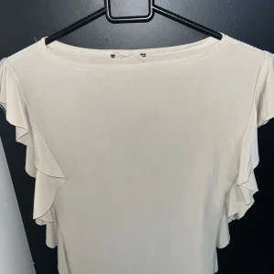 Beige blus med volangärmar - Säljer en stilren beige blus med korta ärmar och snygga volangdetaljer längs axlarna. Toppen har en rak passform och är tillverkad i ett lätt, mjukt material som ger ett luftigt intryck. Perfekt för dig som gillar en enkel men ändå trendig look.