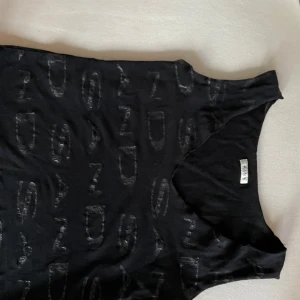 Svart linnetopp från Sand - Svart ärmlös linnetopp från Sand med unikt mönster av gråa bokstäver och siffror över hela plagget. Toppen har rund halsringning och är tillverkad i mjukt material som känns skönt mot huden. Perfekt för dig som gillar en stilren men ändå edgy look.