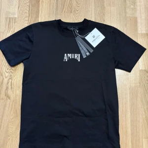 Svart Amiri t-shirt med stor logga - Svart t-shirt från Amiri med stor vit och brun logga på ryggen. Klassisk passform med rund hals och korta ärmar. Perfekt för dig som gillar streetwear och vill sticka ut med en fet logga på ryggen.