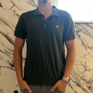 Grön piké från Lyle & Scott - Mörkgrön pikétröja från Lyle & Scott med klassisk krage, tre knappar och gul broderad logga på bröstet. Skön bomullskänsla och korta ärmar, perfekt för en clean och stilren look.