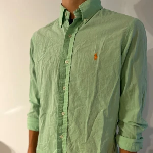 Grön linne skjorta från Polo Ralph Lauren - Snygg ljusgrön linne skjorta från Polo Ralph Lauren i custom fit. Skjortan har klassisk button-down krage, vita knappar och den ikoniska broderade loggan i orange på bröstet. Tillverkad i 100% bomull, perfekt för en fräsch och stilren look.