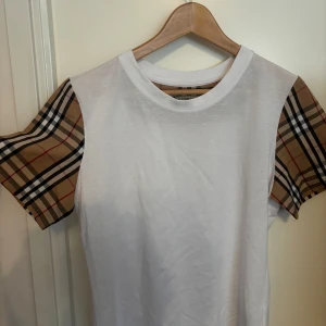 Vit Burberry t-shirt  - Snygg vit t-shirt från Burberry med klassiska beige-rutiga ärmar. T-shirten har rund halsringning och är tillverkad i mjuk bomull. Perfekt för dig som vill sticka ut med en ikonisk detalj från Burberry. Den är M men passar även S.