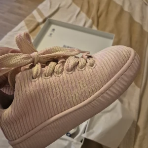 Pastellrosa sneakers från Burberry - Snygga sneakers från Burberry i pastellrosa med stickad textil och breda snören. Använda bara 1 gång då de är för stora för mig . Dustbag och kartong medföljs.    Skorna har en ribbad struktur och platt sula, med deras standard mönstring på                      Nypris var 7900 kr.                                       Säljer de för 3500 kr