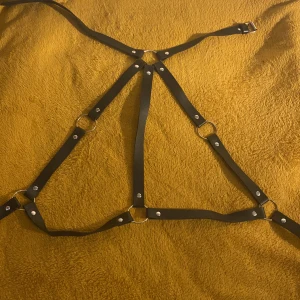 Svart harnessbälte i skinn - Unikt svart harnessbälte i skinn med flera justerbara remmar och silverfärgade metalldetaljer. Bältet har ringar och nitar som ger en edgy vibe och passar perfekt för att lyfta din outfit till nästa nivå. Snygg accessoar för dig som gillar alternativ stil.