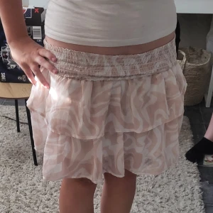 Rosa/ beige/ vit volangkjol med mönster - Supersöt kort volangkjol i beige/ rosa och vitt med abstrakt mönster. Kjolen har bred, smockad midja som är stretchig och två lager volanger i ett tunt, luftigt material. Perfekt för dig som gillar en feminin stil.