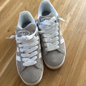 Grå Adidas Campus sneakers mocka - Snygga Adidas Campus sneakers i ljusgrå mocka med klassiska vita ränder och vit sula. Skorna har rund tå, platt sula och breda vita snören. Perfekta för dig som gillar en clean och stilren look med retrovibbar.